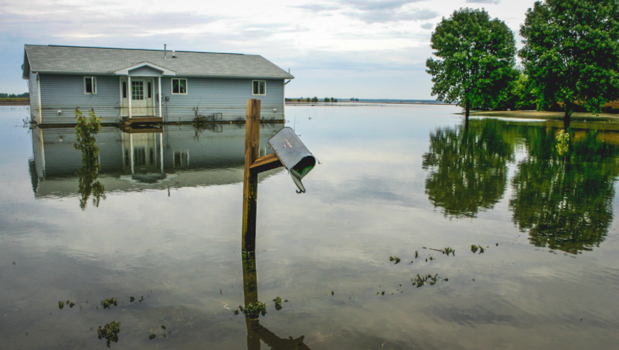 A house sits on a flooded prairie, it's broken mailbox reflected in the mirror-like water.