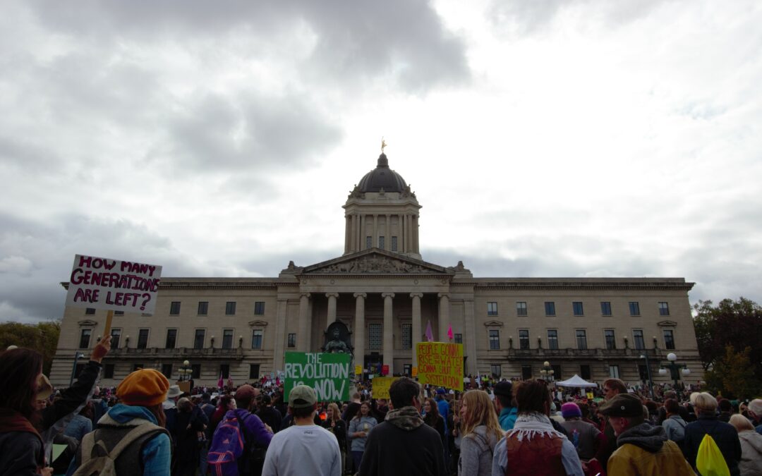 Hundreds of people gathered in front of the Manitoba Legislature for the 2019 General Climate Strike. A few signs are seen above people's head. The most prominent one says "how many generations are left?"