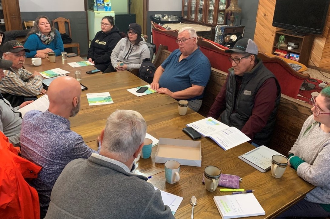 A group of 11 community members sit around a long wooden table facing each other. The table is covered with coffee cups and copies of Manitoba's Road to Resilience.